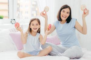 madre e hija divertidas vestidas con ropa de dormir, tienen buen humor por la mañana, toman deliciosos donuts, van a desayunar postre, posan juntos en el dormitorio. concepto de familia y unión foto