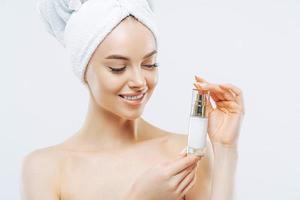 Studio shot of attractive satisfied woman applies moisturizer, holds bottle of skin cream or lotion, wears minimal makeup, poses shirtless against white background, wrapped towel on head. Wellness photo