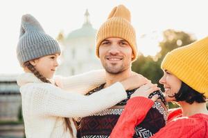 Lovely small girl enjoy time with parents, embrace her father with great love, spend autumn weekends together. Relaxed young people in knitted hats have happy expressions. Togetherness concept photo