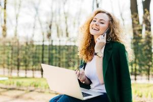 Portrait of beautiful girl having fluffy blonde hair holding smartphone in one hand and sunglasses in other having telephone conversation being excited to speak with her best friend using laptop photo