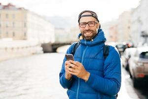 Happy man with beard wearing eyeglasses dressed in blue anorak holding backpack and mobile having happy look travelling in city watching sightseeings and making photos having exursion outdoor