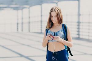 Outdoor shot of pleasant looking Caucasian female with slim body, carries rucksack, holds modern cell phone, listens radio online with earphones, reads notification, searches internet websites photo