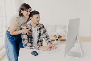 Caring young happy woman leans at husbands shoulders, worries about him, focused into computer monitor, keyboard and surf internet, pose at workplace in modern apartment, little dog sits near photo