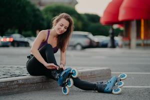 People leisure fitness sport recreation concept. Pleased young woman puts on rollerskates going to ride rolles in urban place has regular exercising goes in for dangerous sport adjustes laces photo