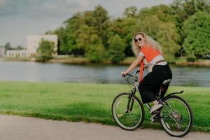 Photo of cheerful woman dressed casually, rides bicycle, looks aside, has happy expression, wears shades, poses near river, green lawn and trees, some buildings in background. People and rest
