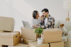 esposo y esposa felices se mudan a una casa nueva, compran muebles en línea, usan tecnología moderna, el hombre navega por Internet a través de una computadora portátil y habla por teléfono, se sienta en el sofá con su novia y su perro, muchas cajas de cartón foto