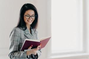 Satisfied brunette woman has happy expression writes down working plan in notepad has red manicure formal clothes records information for organization process wears spectacles notes financial schedule photo