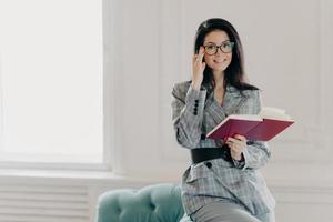 Smiling female business reporter in formal clothes holds opened notebook and pen, writes down information or checklist, wears formal clothes, works remotely from home, prepares for formal meeting photo