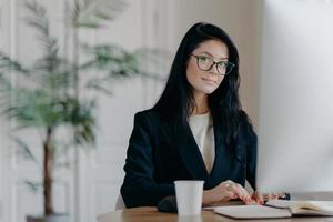 Serious successful female office worker in formal outfit, wears eyeglasses, busy with online work, poses at desktop, works on computer, reads business new or watches tutorial, enjoys working process. photo