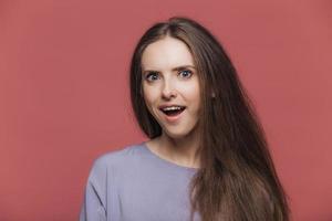 Amazed satisfied beautiful female model with dark straight hair, keeps mouth opened, hears unexpected news, poses against pink studio background. People, emotions, facial expressions, feelings concept photo