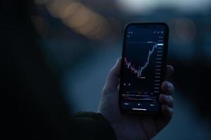 Trader holding smartphone with financial chart and analyzing price flow on stock market photo