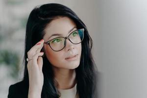 Close up shot of brunette serious lady keeps hand on rim of spectacles, concentrated at computer screen, watches online video, poses indoor, browses web page, creats project, poses in office photo