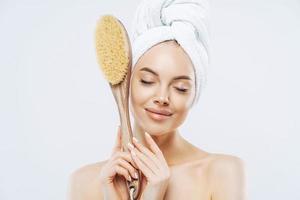 toma horizontal de mujeres complacidas con los ojos cerrados, sostiene un gran cepillo de baño para masajear la piel, disfruta de tratamientos higiénicos en casa, aislada en fondo blanco. belleza, cuidado, exfoliación foto