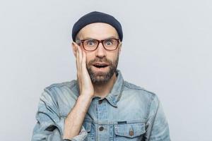 Portrait of amazed bearded excited male looks with surprisment in camera, recieves unexpected news, dressed in fashionable clothing, has attractive look, isolated over white studio background photo