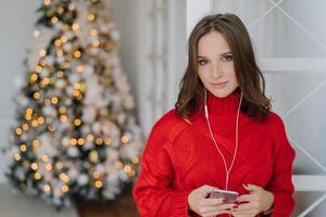 foto de una linda mujer joven con cabello oscuro, vestida con un suéter rojo de punto, sostiene un teléfono inteligente en las manos, escucha radio en línea, está conectada a wifi en casa, posa cerca del hermoso árbol de año nuevo con luces