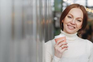 una foto de una mujer atractiva con una sonrisa dentuda, maquillada, vestida con un suéter blanco de cuello alto con cuello, toma café para llevar, disfruta del tiempo libre, expresa buenas emociones. concepto de personas y ocio