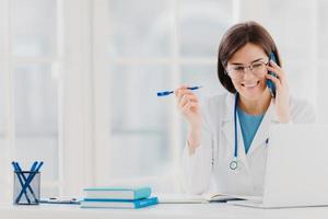 Glad professional doctor concentrated in modern laptop computer, reads useful information, has telephone conversation, discusses medical issues, sits at hospital office with notepads on table photo