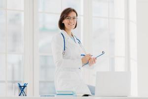 Indoor shot of professional female doctor supplies medic care assistance, poses at desktop with laptop, prepares document agreement paper offer, wears optical glasses and white medical gown. photo