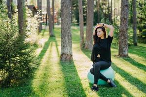 Fitness woman looks into distance with thoughtful expression sits on fitness ball wears black t shirt and leggings poses at green spring forest, keeps arms raised over head, has workout outdoor photo