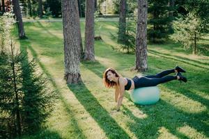 Above view of determined young brunette European woman exercises pilates in green park, balances on fitness ball, dressed in activewear, looks happily somewhere poses outside. Selective focus photo