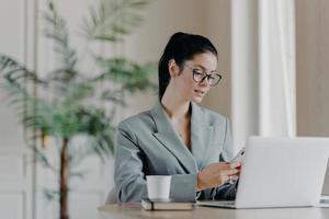 Gorgeous busineswoman types message on smartphone, works on new publication, poses in front of laptop computer, involved in working process, sits at desktop, drinks aromatic coffee, uses internet photo