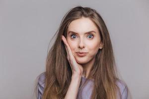 Attractive blue eyed brunette female looks with bewilderment at camera, has specific appearance, cant beleive in eyes, isolated over grey studio background. People and facial expressions concept photo