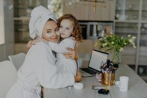 una mujer atractiva usa parches de hidrogel debajo de los ojos vestidos con albornoz y una toalla envuelta abraza a su pequeña hija con pose de amor juntos en casa cerca de herramientas cosméticas. concepto de rutina de maquillaje foto