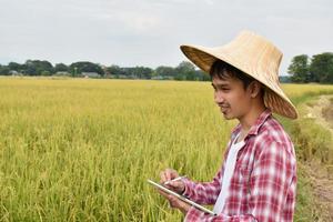 Portrait of Asian younger farmer who wears hat and red-white shirt, holds taplet, stands in the middle of rice paddy field, concept for using technology with agriculture in dailylife. photo