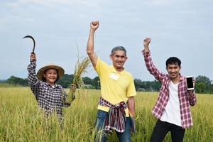 tres agricultores asiáticos están parados en el campo de arroz y levantando la mano para felicitar y regocijarse por la finalización exitosa de la encuesta de cultivo de arroz y el almacenamiento de datos mediante el uso de teléfonos móviles. foto