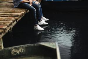 Cropped photo young lovers married couple, husband and wife, held of hands sit on a wooden bridge near lake. lower half. Place for text.