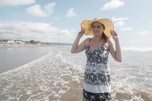 Beautiful happy young Hispanic woman walking alone on the beach wearing a hat and black and white dress during a sunny morning photo