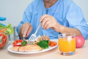 Asian senior or elderly old lady woman patient eating Salmon steak breakfast with vegetable healthy food while sitting and hungry on bed in hospital. photo