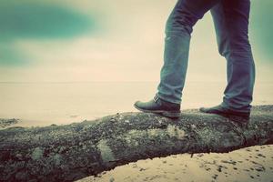 Man in jeans and elegant shoes standing on fallen tree on wild beach looking at sea. Vintage photo