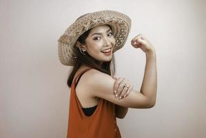 Gorgeous strong young Asian woman with orange dress and straw hat showing biceps and smiling. Indonesian girl strong concept. photo