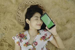 A young beautiful Asian woman lying on the sand while showing phone screen photo