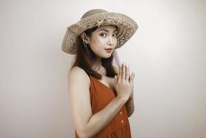 A young beautiful Asian woman wearing a straw hat gives greeting hands with a big smile on her face. Indonesian woman on white background. photo