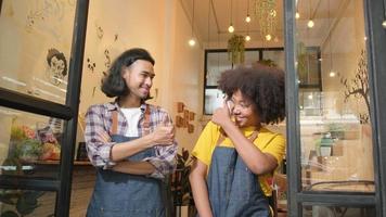 Two young startup barista partners with aprons stand at casual cafe door, arms crossed, laugh and tease together, happy and cheerful smiles with coffee shop service jobs, small business entrepreneurs. video