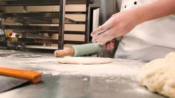 câmera lenta e close-up, chef em uniformes de cozinheiro branco com aventais polvilhe farinha, amassar massa de pastelaria com rolo, preparar pão, tortas e comida de padaria fresca na cozinha de aço inoxidável do restaurante. video