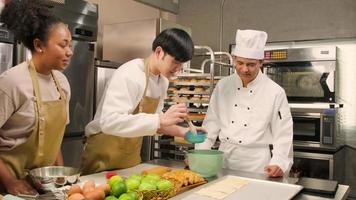 curso de culinária, chef masculino sênior de uniforme ensina jovens estudantes de aula de culinária, escova a massa com creme de ovos, prepara ingredientes para alimentos de padaria, tortas de frutas na cozinha de aço inoxidável. video