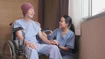 Uniformed young Asian female therapy doctor holding hands wheelchair male patient at window to support and motivate recovery, cancer illness after chemo medical treatment in hospital inpatient room. video