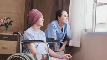 Uniformed young Asian female therapy doctor encouraging wheelchair male patient at window to support and motivate recovery, cancer illness after chemo medical treatment in hospital inpatient room. video