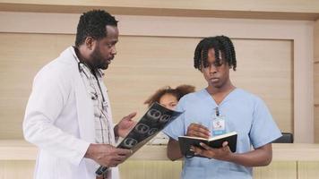 il medico maschio afroamericano professionista discute con uno studente di medicina e un assistente sulla diagnosi di un film radiografico e sulla medicina di un paziente al bancone della reception dell'ambulatorio dell'ospedale. video