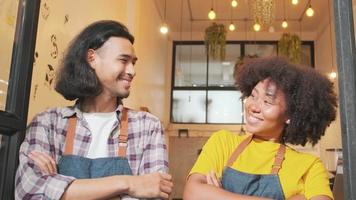 två unga startup-baristapartners med förkläden står vid casual kafédörren, armarna i kors, skratta och retas tillsammans, glada och glada leenden med kaféservicejobb, småföretagare. video