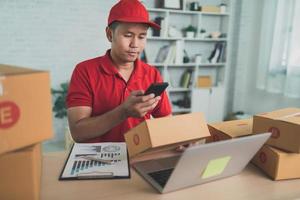 Asian delivery man worker smiling using mobile scan barcode for boxes for sending or conveying parcels by mail. Wearing red uniform work at home. photo