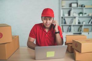 Asian delivery man worker smiling calling mobile phone to customers for boxes for sending or conveying parcels by mail. Wearing red uniform work at home. photo