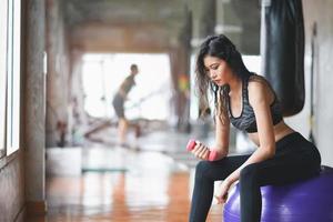 mujer deportiva en forma asiática sentada en la pelota y haciendo ejercicio con pesas en la sala de pesas en el gimnasio. concepto de fitness de mujer deportiva foto