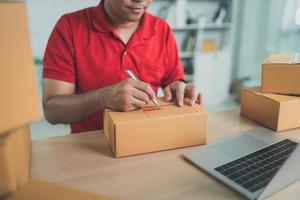 Asian delivery man worker in red uniform work at home , checking list parcel boxes for sending or conveying parcels by mail. photo