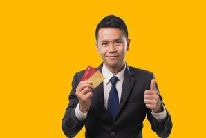 Asian businessman holding red credit card and mobile phone while smiling and online shopping isolated yellow background. Asian man buy something with credit card shopping online concept. photo