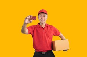 un repartidor asiático sonriendo y sosteniendo una tarjeta de crédito con uniforme rojo aislado en un fondo amarillo, sosteniendo cajas de paquetes para enviar o transportar paquetes por correo. foto