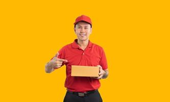 trabajador asiático de entrega sonriendo y señalando con el dedo la caja con uniforme rojo aislado en fondo amarillo, sostenga cajas de paquetes para enviar o transportar paquetes por correo. foto